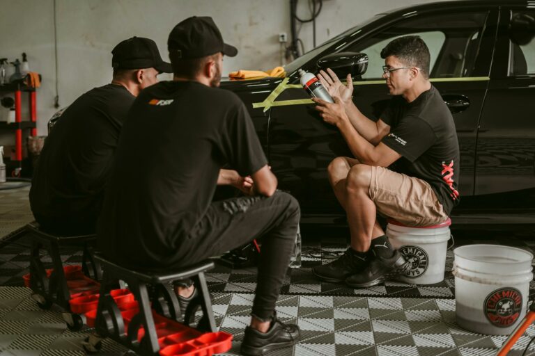 Men discussing car detailing techniques in a professional workshop setting.